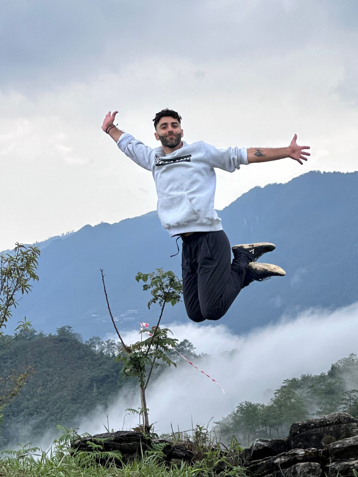 Cava jumping with arms open above a misty mountain valley